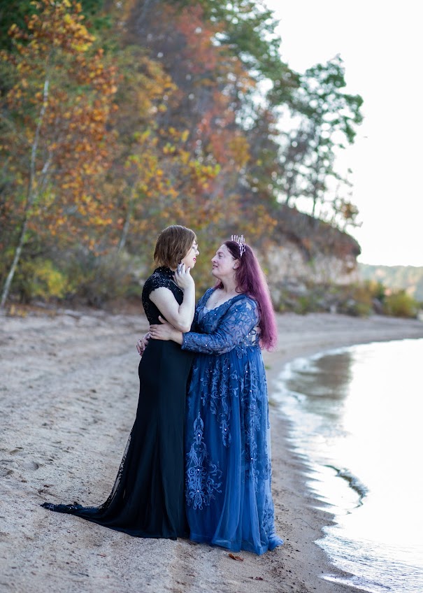Photograph by Denise Cerniglia. The picture shows two women in lacy floor-length gowns standing by the shore of a lake near dusk. The woman on the left is slim and has light skin, short brown hair, and glasses. She is wearing a short-sleeved, high-necked black gown with a small train. The woman on the right is heavier, with light skin and long reddish-purple hair. She is wearing a lacy blue dress with a low neckline. On her head is a small silver headpiece or tiara. Her arms are around the other woman's waist. The women are looking at each other and look happy. In the background is a wooded hillside. The leaves on the trees are beginning to turn yellow and red.